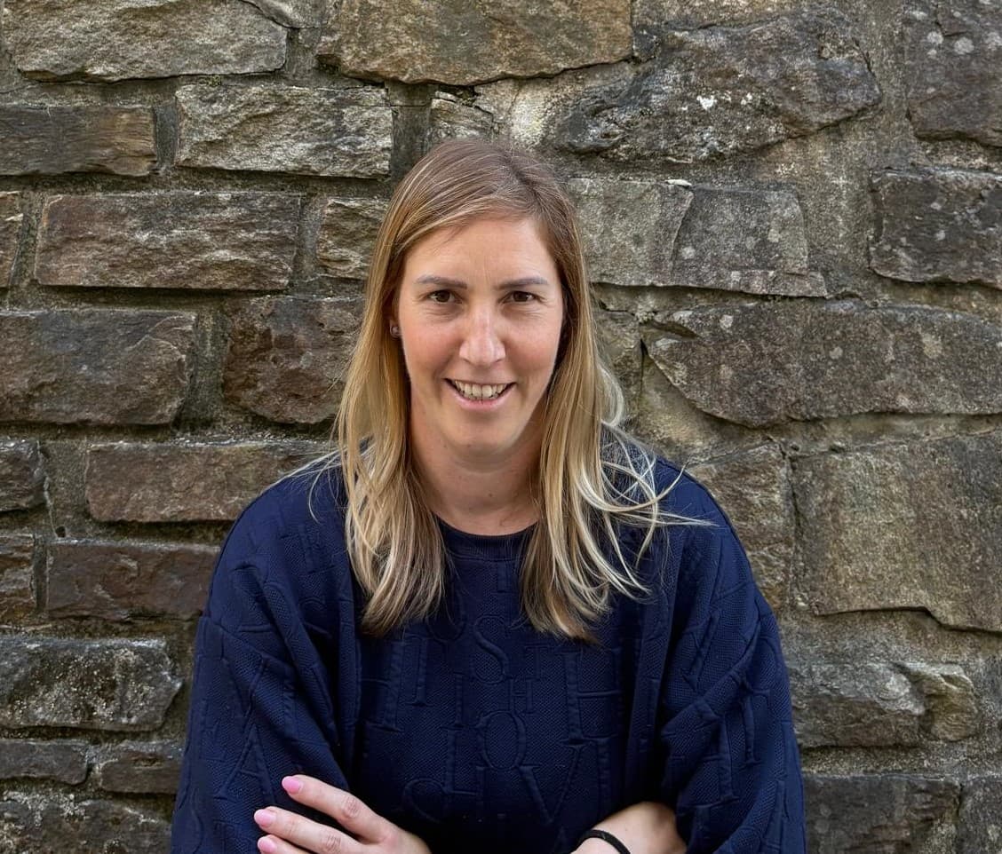 Smiling blonde woman wearing a navy blue sweater posing against a rustic stone wall.