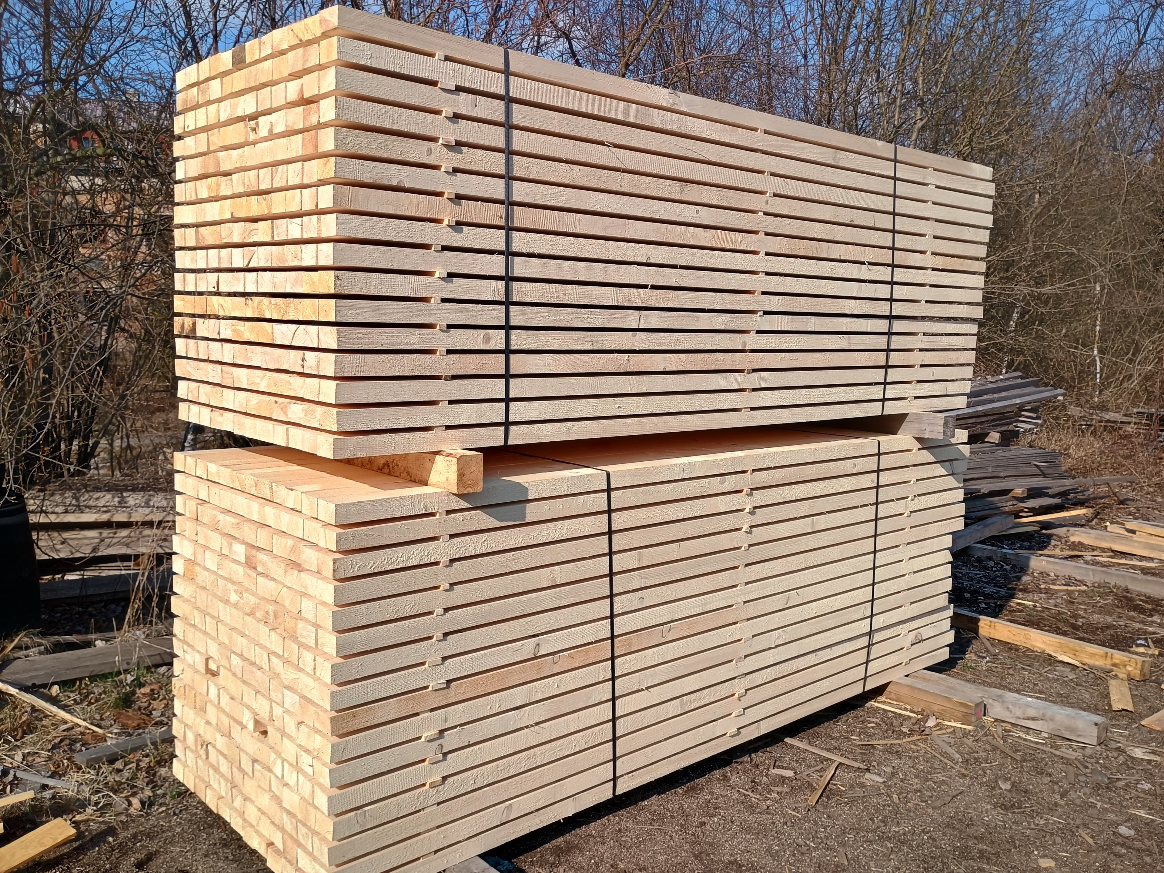 Two large stacks of wooden planks bundled with black straps in a sunny outdoor yard.