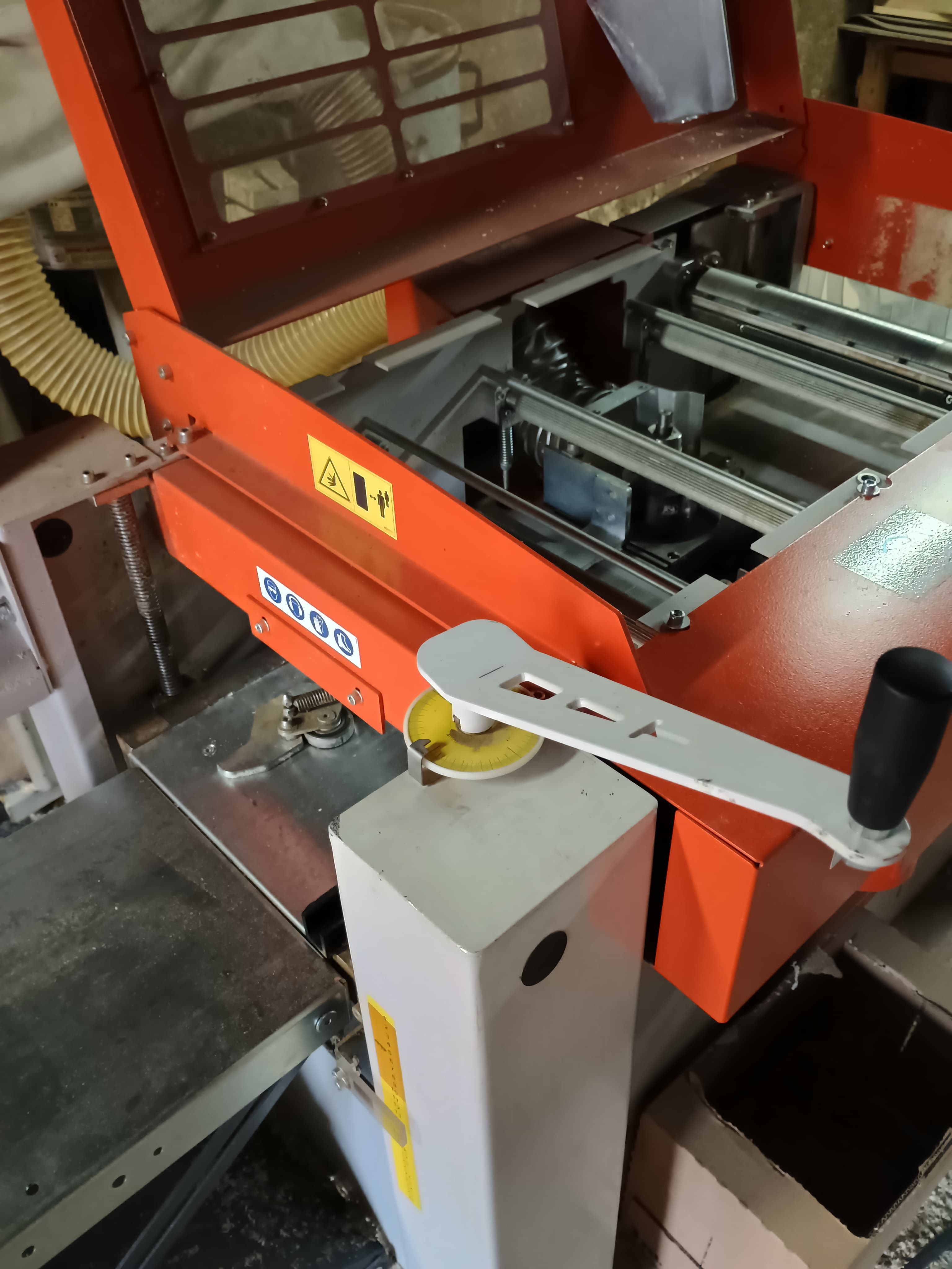 Industrial orange machine with a white hand crank, safety stickers, and yellow flexible hose.