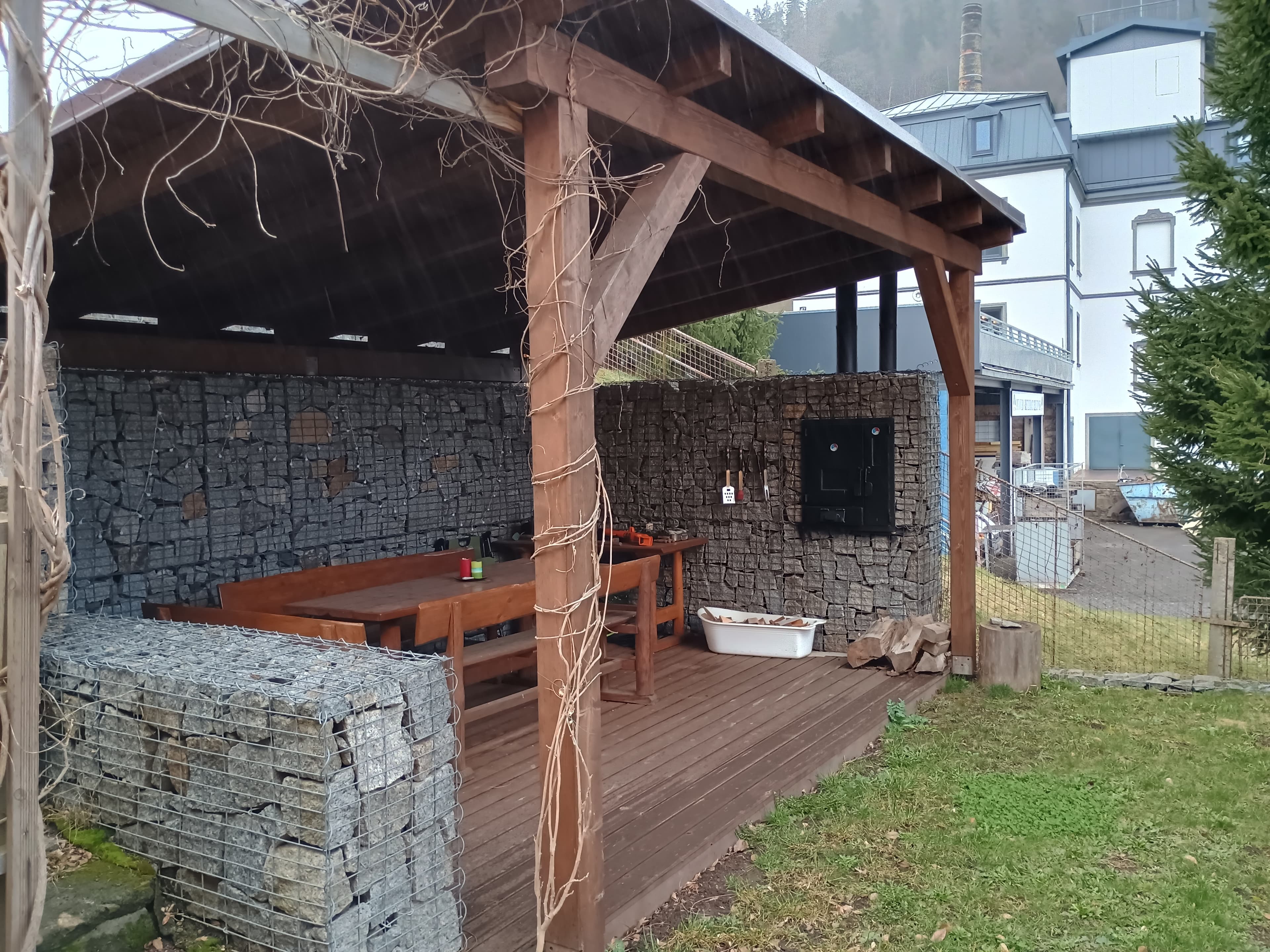 Outdoor wooden gazebo with gabion stone walls, a dining table, and a built-in oven.