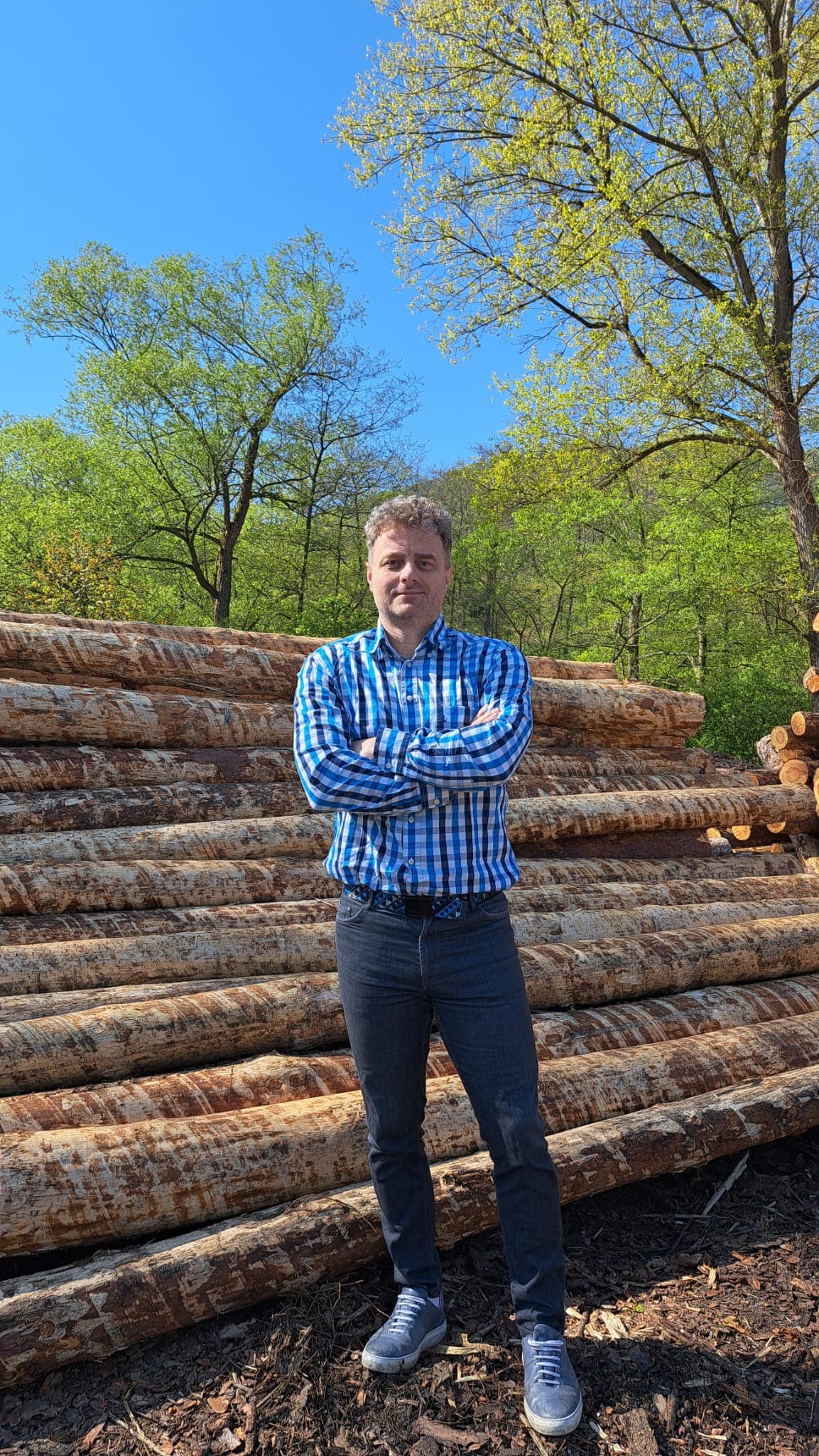 Man in blue checkered shirt stands with arms crossed before a large stack of logs.